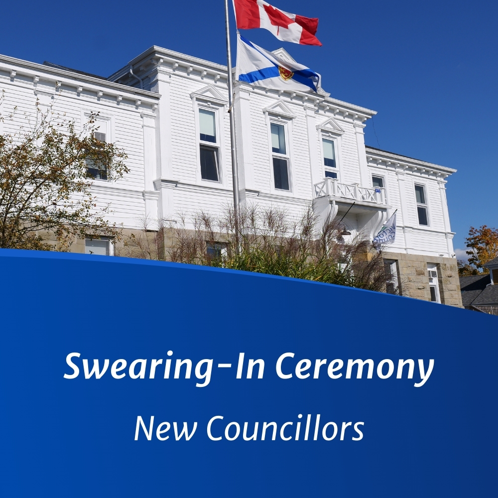 Photograph of the exterior of the Victoria County Municipal Building, with the Canadian and Nova Scotia flags waving in the breeze against a blue sky with text reading "Swearing-In Ceremony - New Councillors