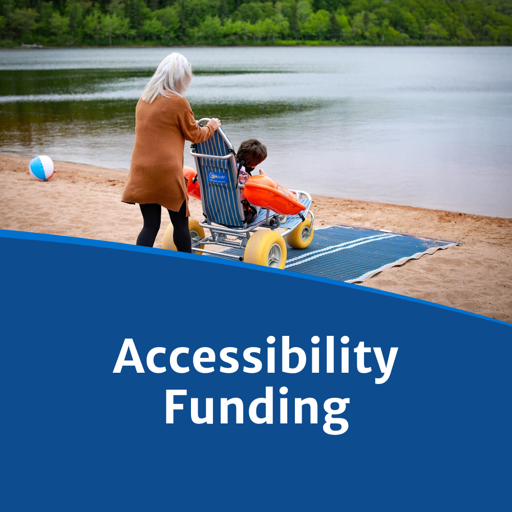 A woman pushes a person in a wheel chair on an access mat leading towards the water on a beach.