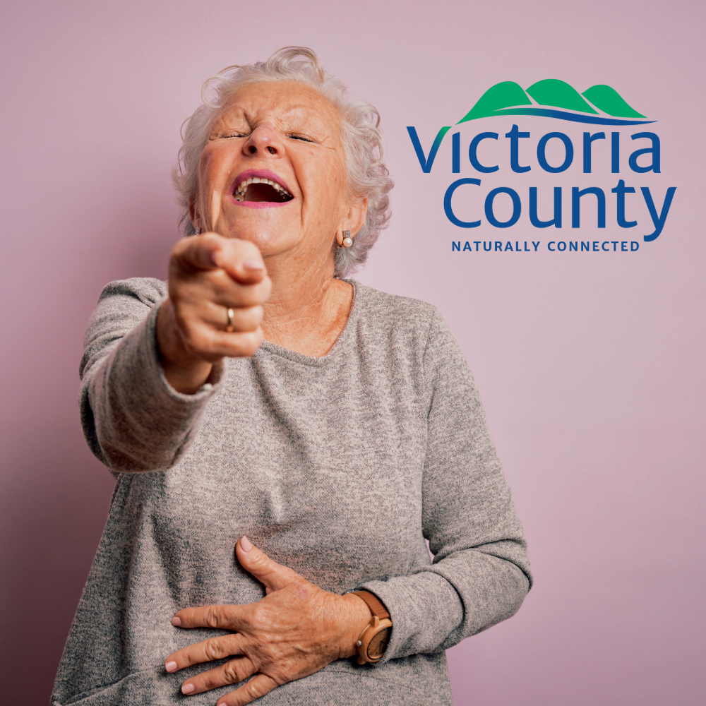 A woman with short grey hair is laughing and pointing at the camera.